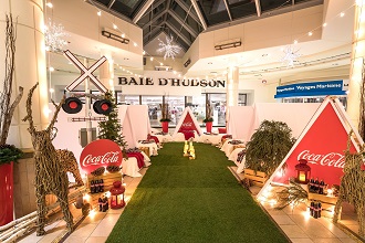 Installation des Fêtes devant La Baie d’Hudson avec un sentier de gazon synthétique vert, des rennes en osier, des arbres illuminés et des enseignes rouges Coca-Cola.
