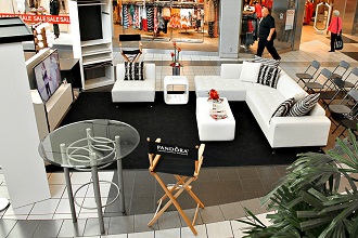 Aménagement de salon dans un centre commercial avec des canapés sectionnels blancs sur un tapis noir, une table en verre et une chaise de réalisateur portant la marque Pandora.