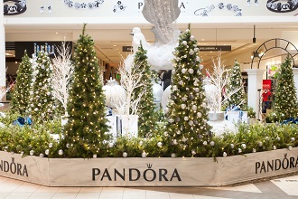 Décor des Fêtes dans l’allée d’un centre commercial avec des sapins de Noël illuminés, des ornements blancs et une sculpture de cygne dans une jardinière incurvée portant la marque Pandora.
